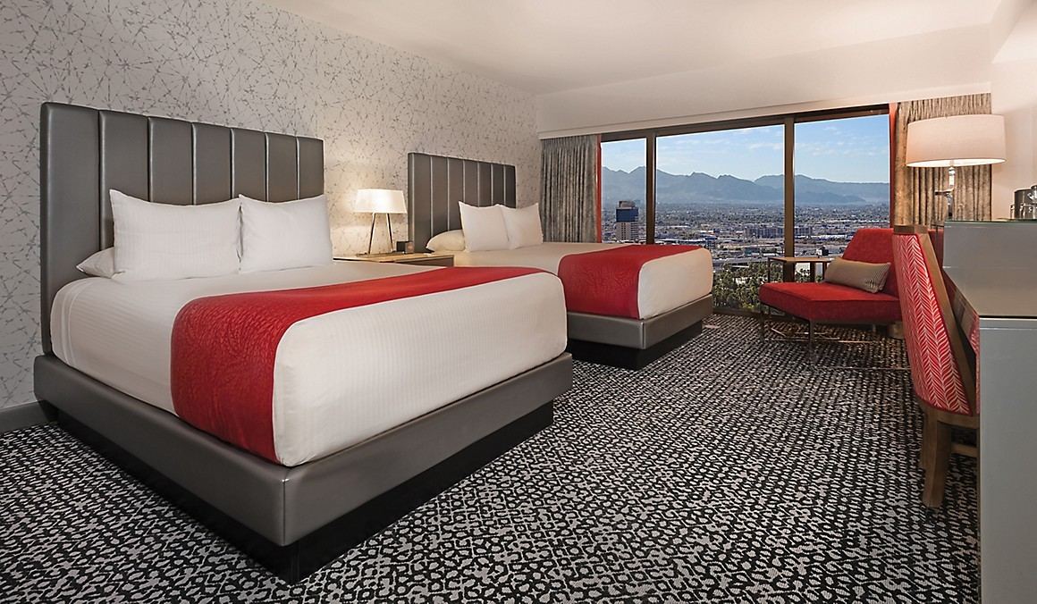 Hotel room with two beds, red runners, patterned carpet and city-to-mountain window view